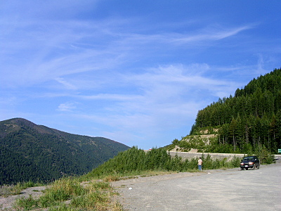 [An expanse of blue sky with a mountain on the right with a road curving around it and a paved area to pull off it. There is another mountain on the left covered with evergreens.]